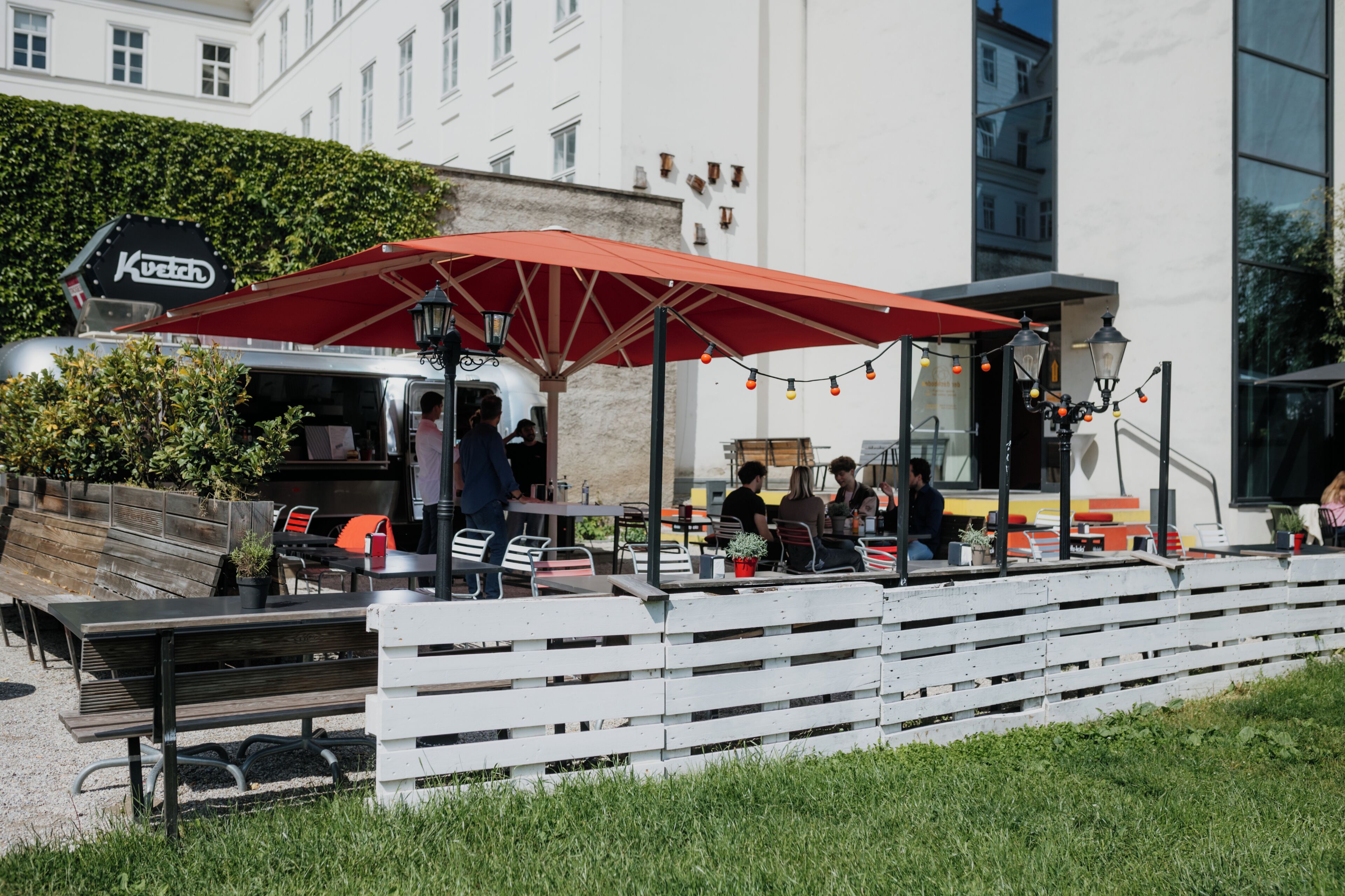 Um café ao ar livre com mesas e cadeiras, um guarda-sol vermelho e um letreiro que diz 'Küstel'.