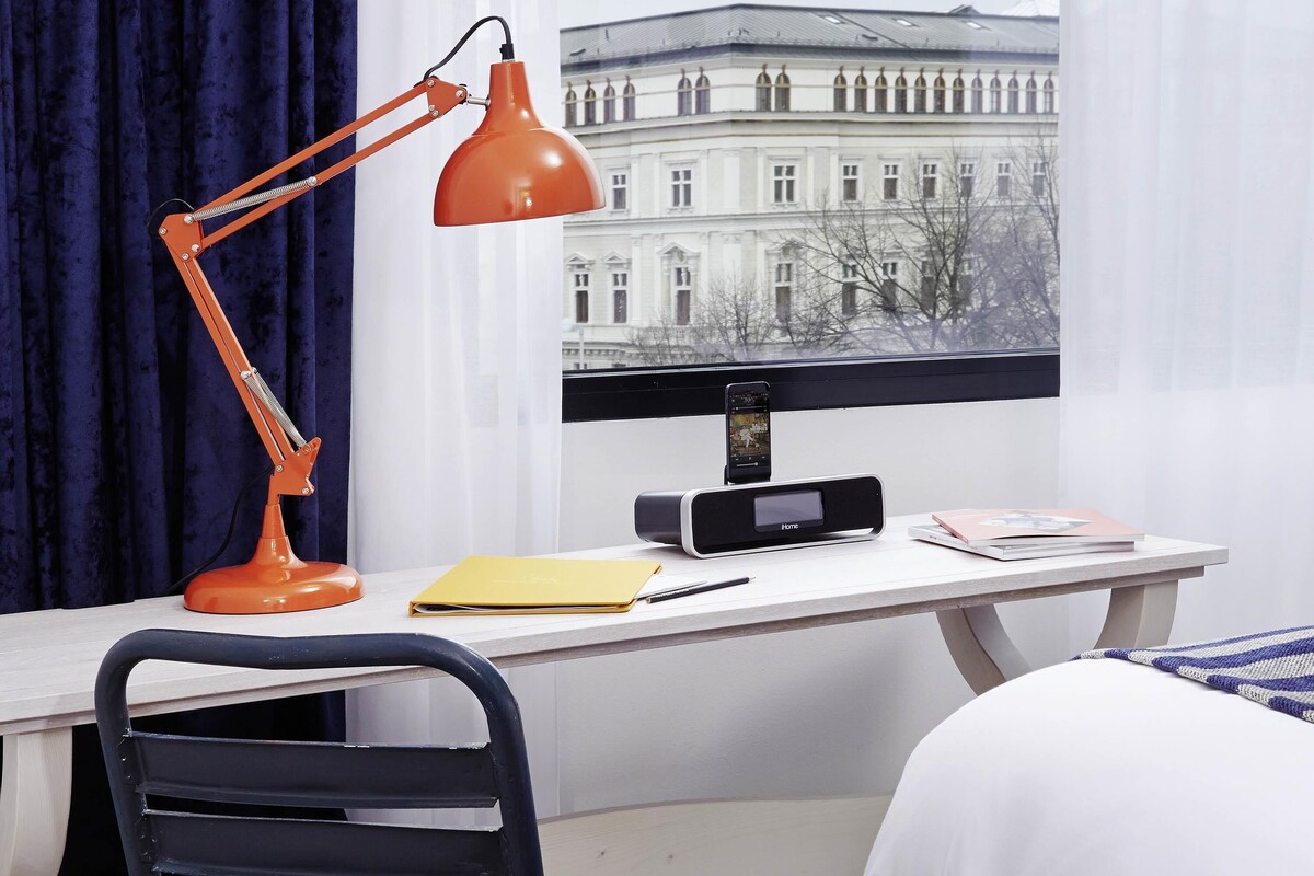 A hotel room with a desk, a black chair, a red desk lamp, a black radio, a yellow folder, a black smartphone, and a bed with white bedding.