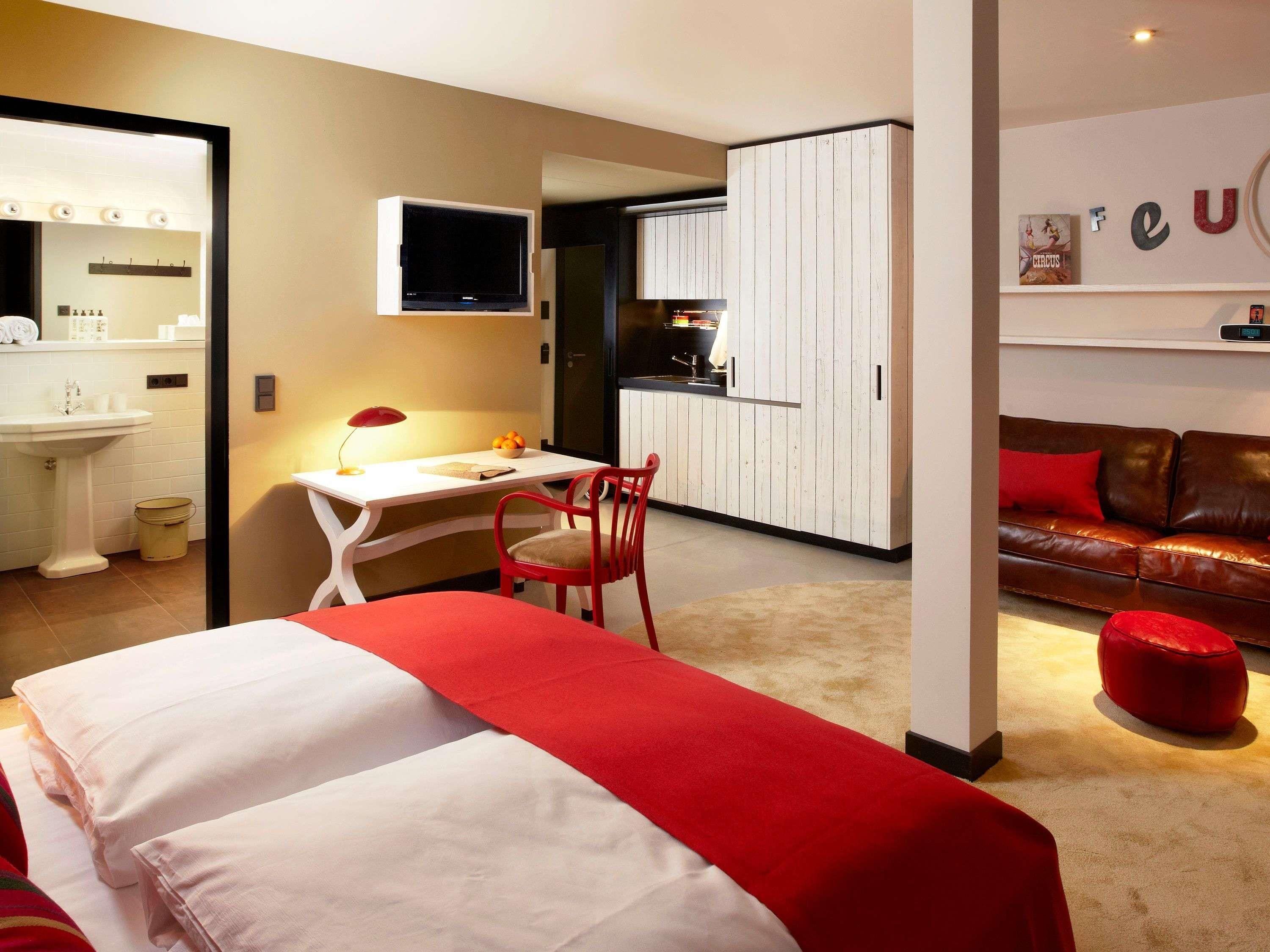 Une chambre d’hôtel avec un lit, un bureau, une chaise, un pouf rouge et un canapé rouge.