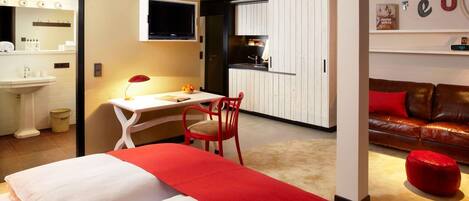 Une chambre d’hôtel avec un lit, un bureau, une chaise, un pouf rouge et un canapé rouge.
