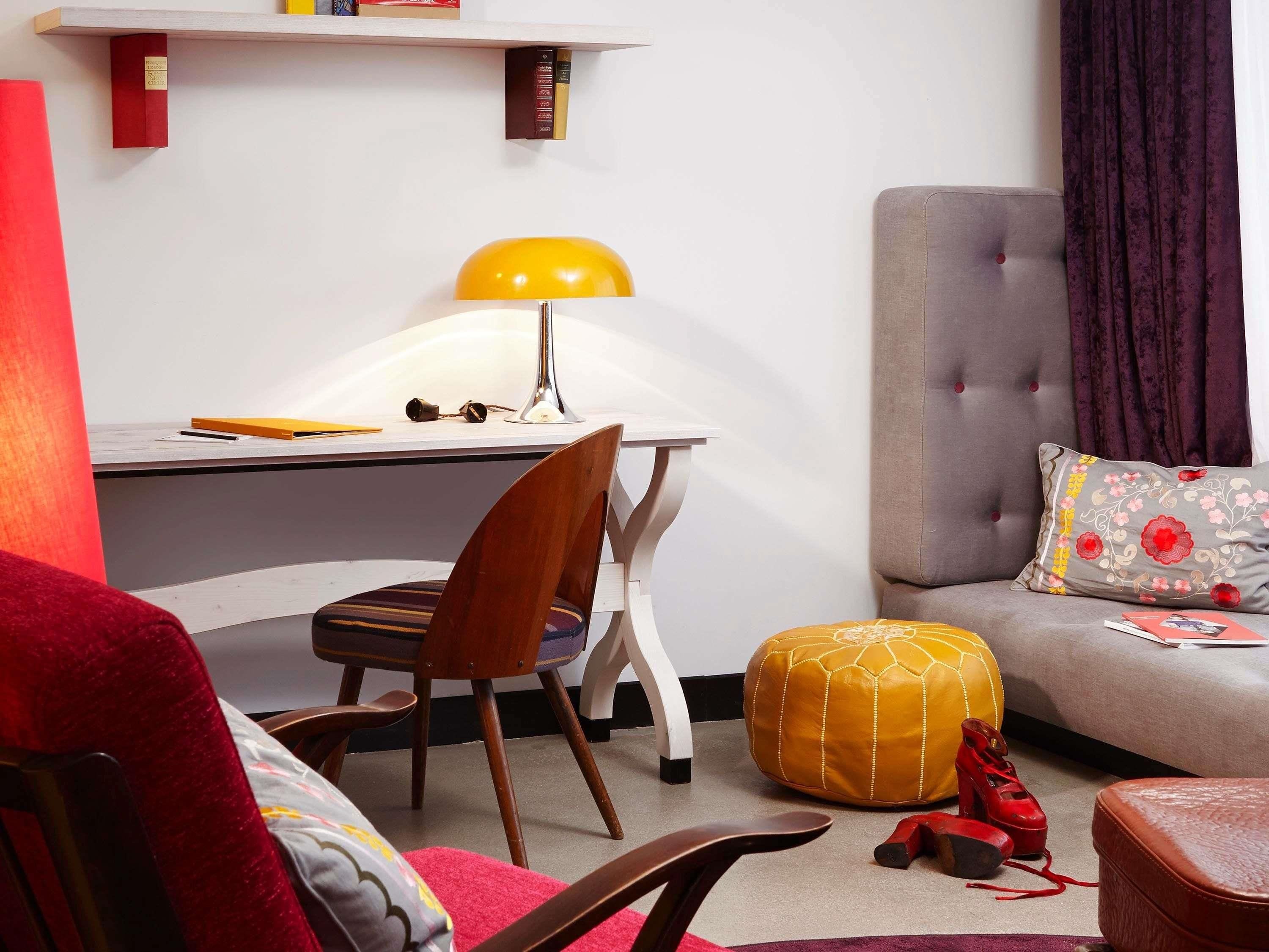 Una sala de estar moderna con un escritorio blanco, una lámpara amarilla, un sofá gris con un cojín estampado y una silla roja.