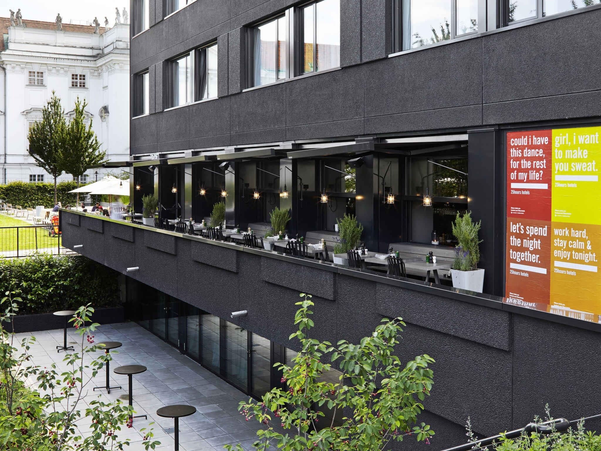 Un espacio exterior para comer con mesas y sillas, fachada de un edificio moderno y un cartel con texto.