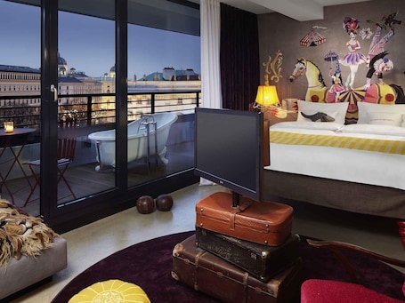 A hotel room with a large window, a television, a bed, a wall mural, and a balcony with a view of a cityscape.. 25hours Hotel Vienna at MuseumsQuartier