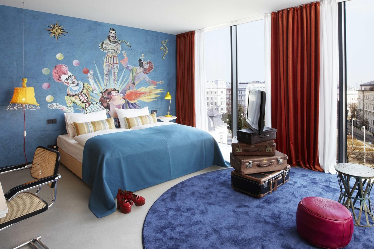 A hotel room with a bed, a TV, a red ottoman, a round blue rug, and a city view.