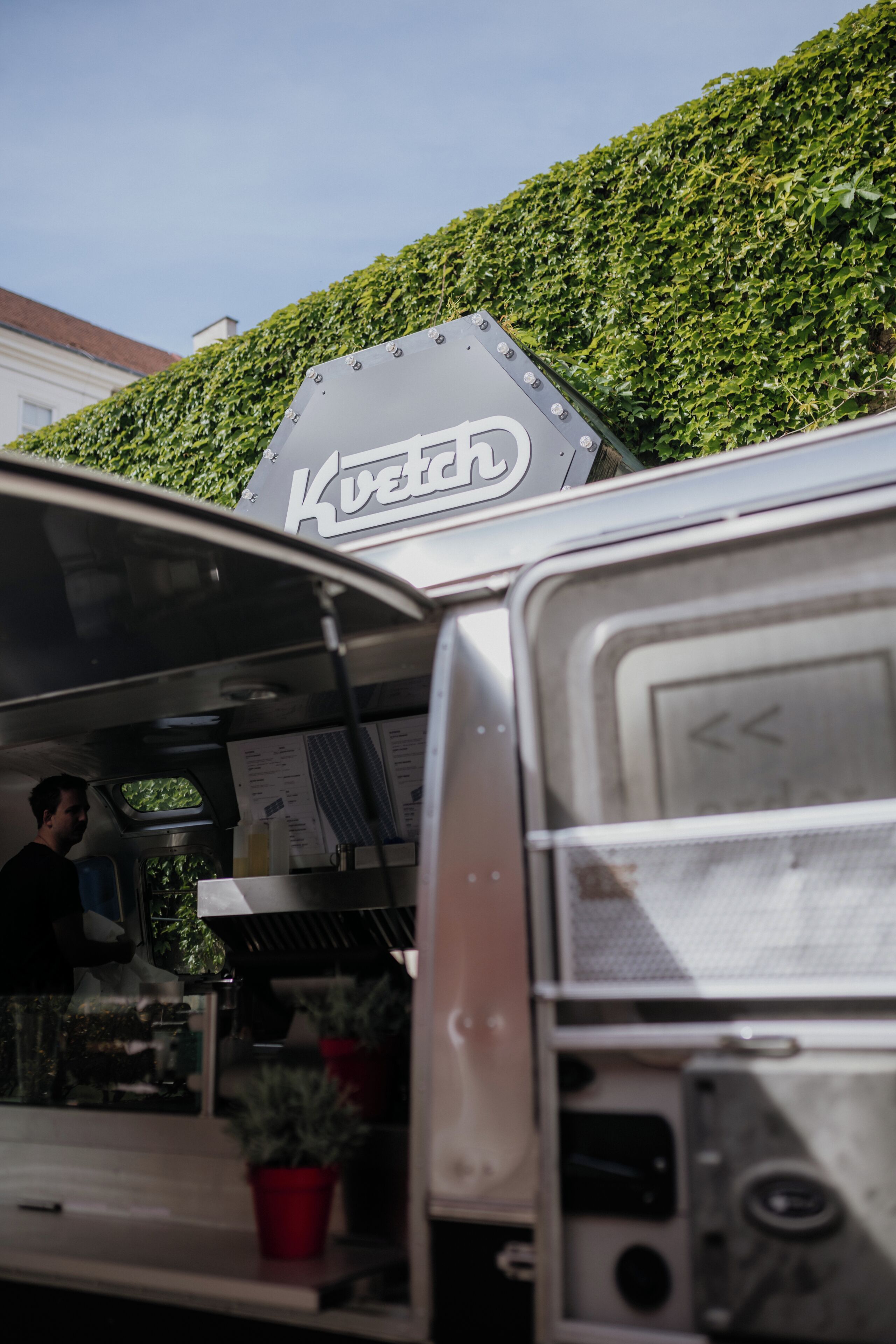 A food truck with a 'Kvitah' sign, a person inside, and a green hedge in the background.