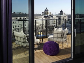 A balcony with a view of historic buildings, a table with two glasses, and a purple pouf. - 25hours Hotel Vienna at MuseumsQuartier (Vienna)
