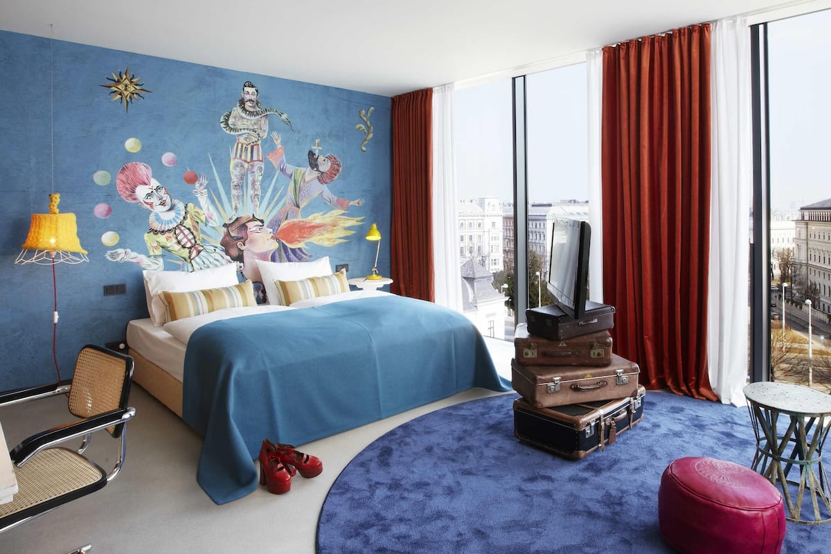 A hotel room with a bed, a TV, a red ottoman, a round blue rug, and a city view.