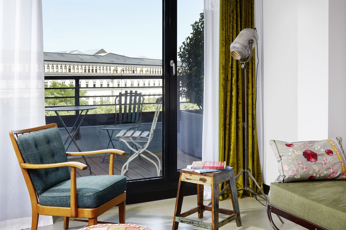 A room with a view of a building, a chair, a table, a lamp, and a sofa.