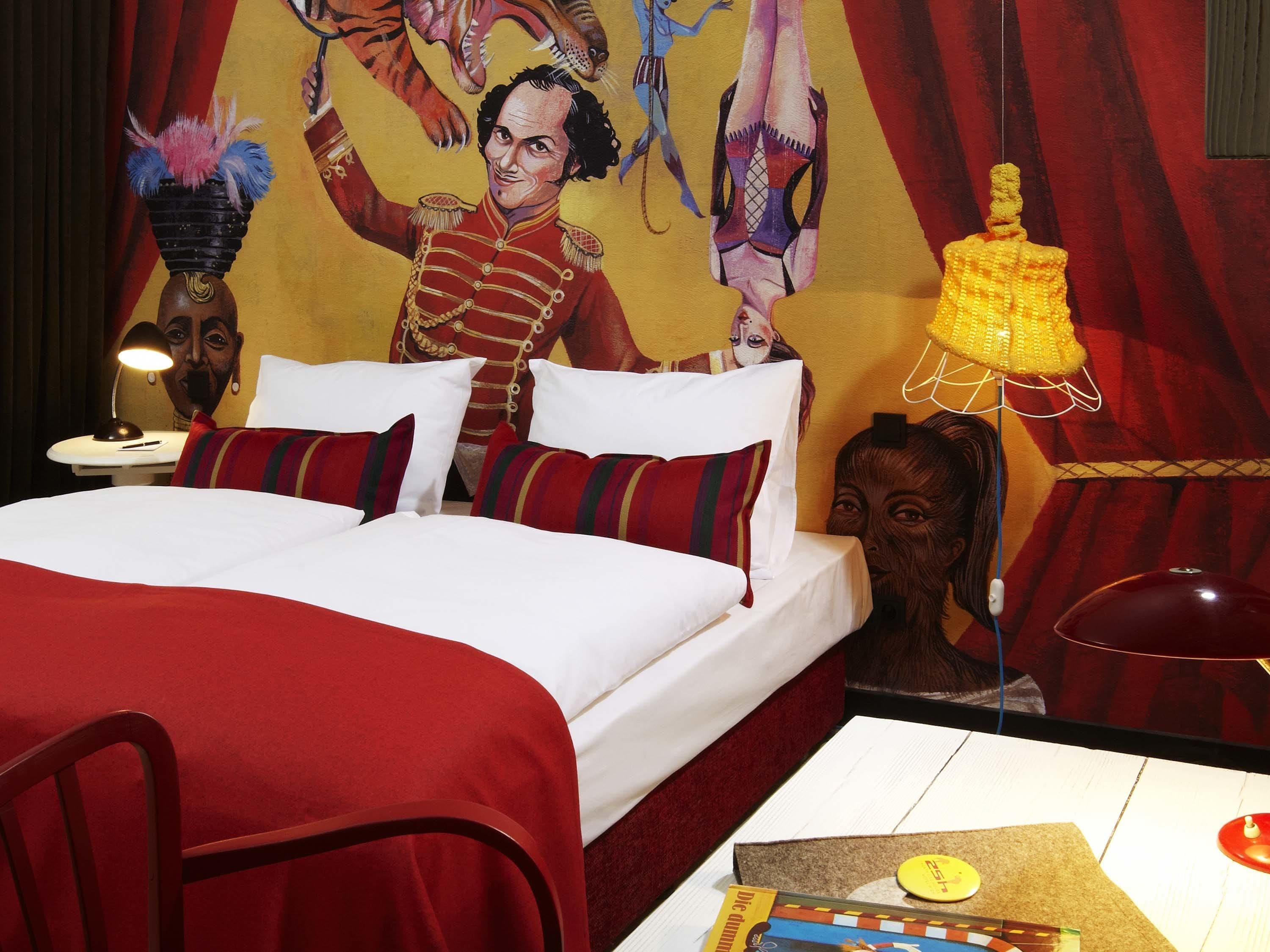 Quarto de hotel com cama, almofadas, mesa de cabeceira, luminária, painel decorativo na parede e uma cadeira.