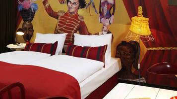 Quarto de hotel com cama, almofadas, mesa de cabeceira, luminária, painel decorativo na parede e uma cadeira.