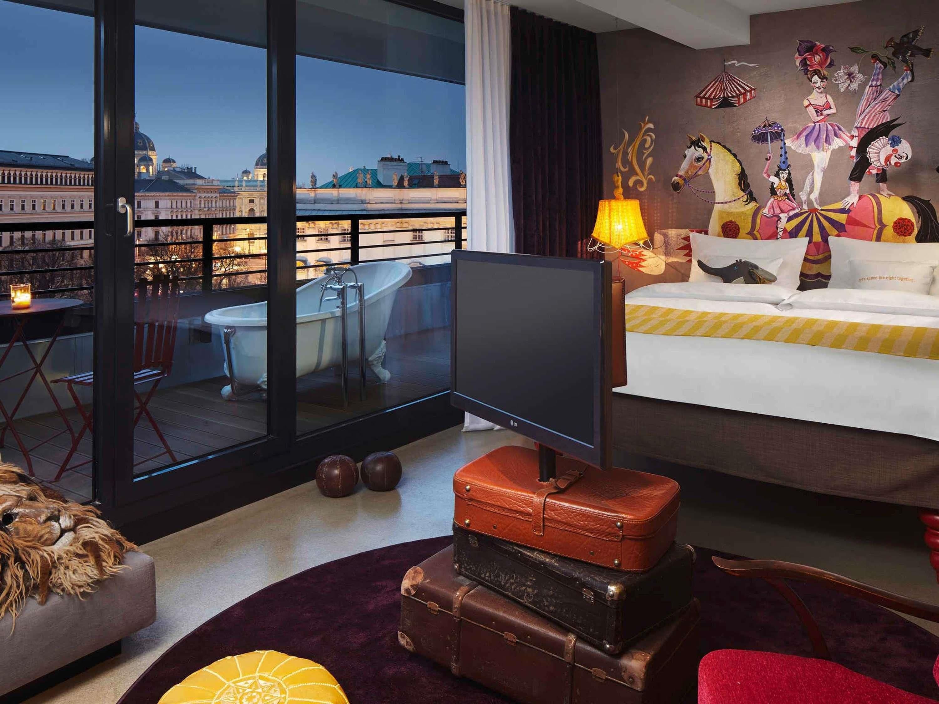 Habitación de hotel con un ventanal grande, un televisor de pantalla plana, una cama, un mural en la pared y un balcón con vistas a un paisaje urbano.