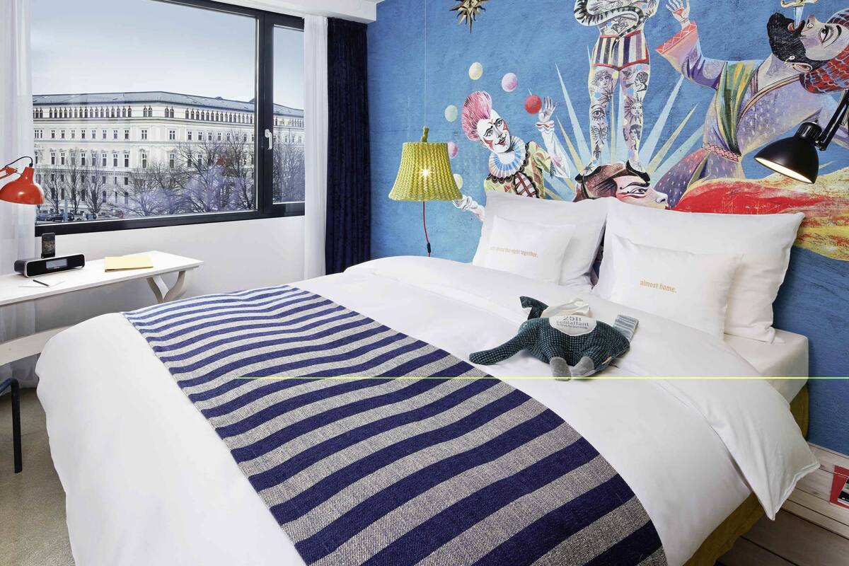A hotel room with a large bed, a desk with a lamp, a view of a building through a window, and a colorful mural on the wall.