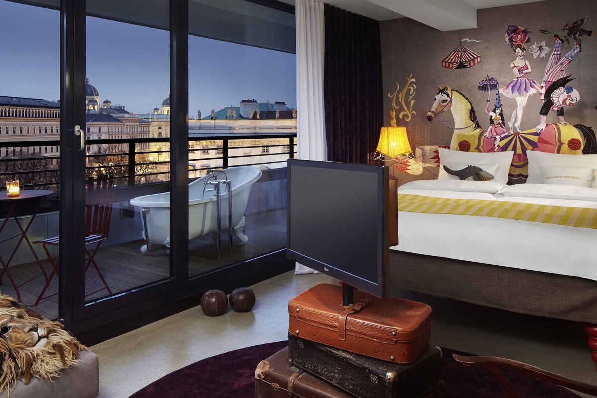 A hotel room with a large window, a television, a bed, a wall mural, and a balcony with a view of a cityscape.