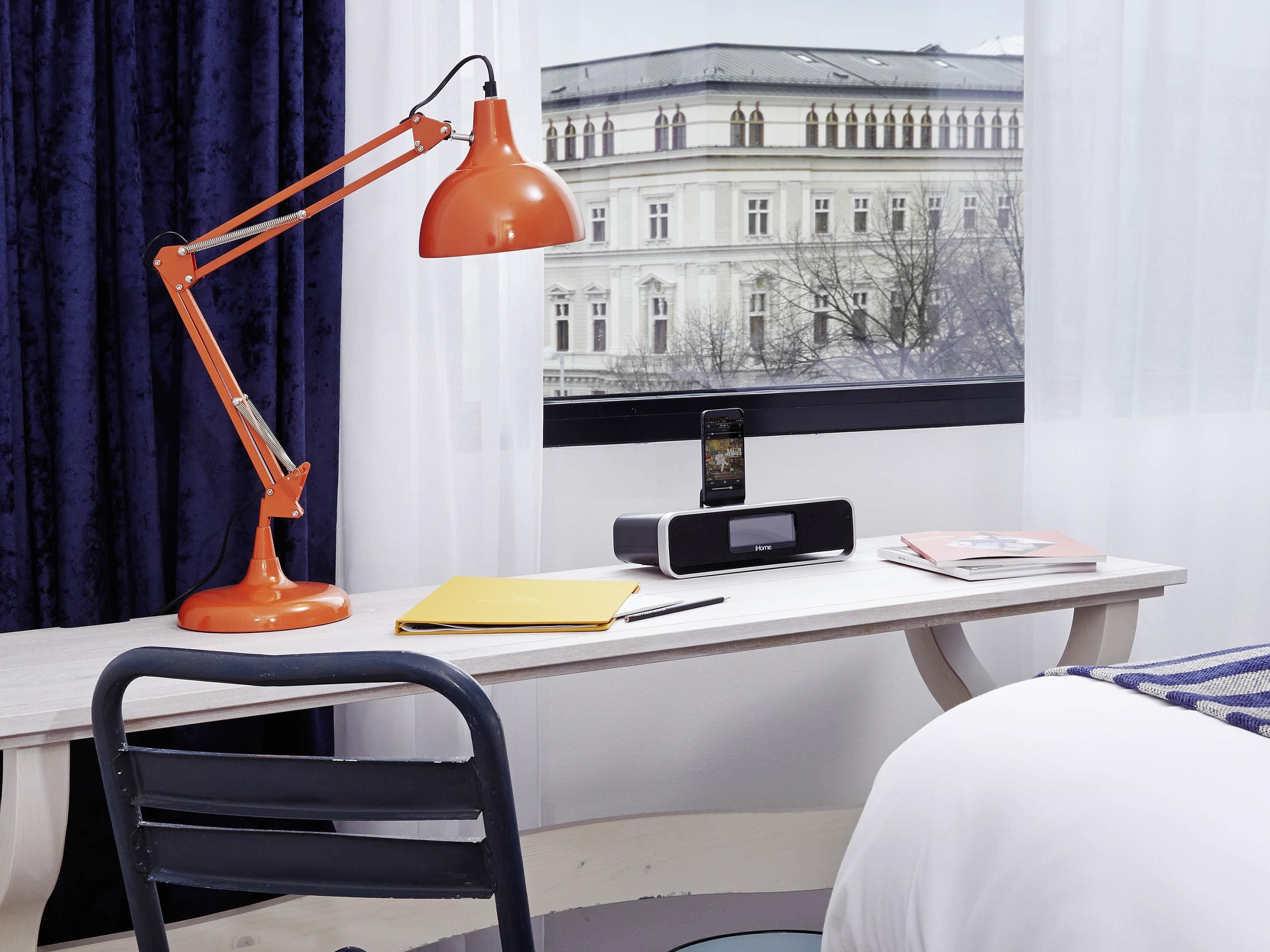 Hotelkamer met een bureau, een zwarte stoel, een rode bureaulamp, een zwarte radio, een geel dossier, een zwarte smartphone en een bed met wit beddengoed.