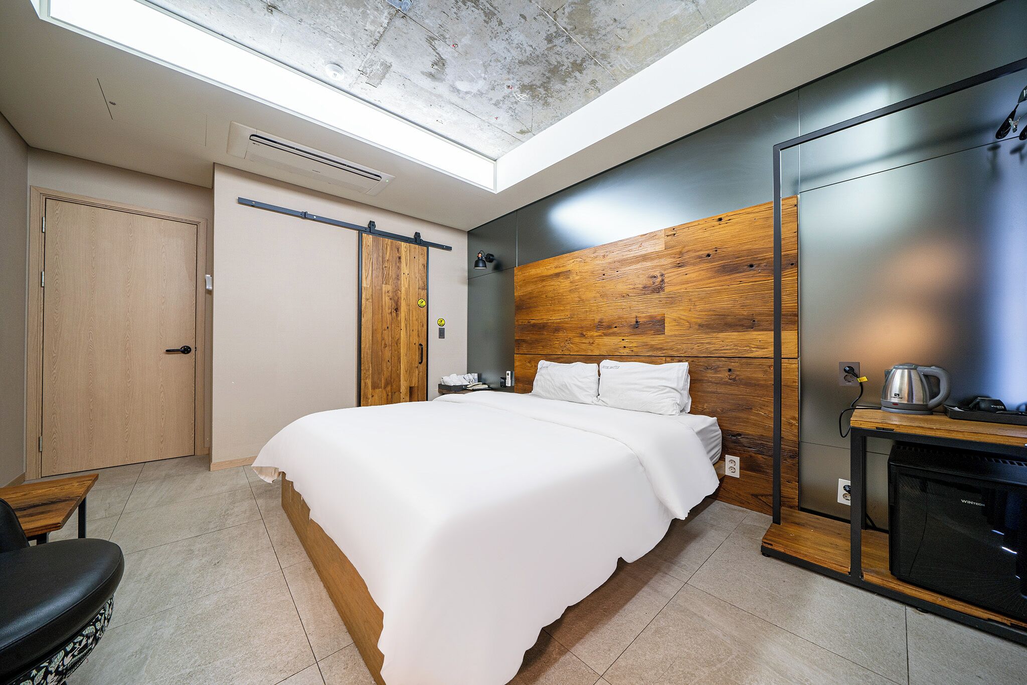 Un dormitorio moderno con tragaluz, cabecera de madera, cama con ropa blanca, escritorio con silla y horno empotrado.