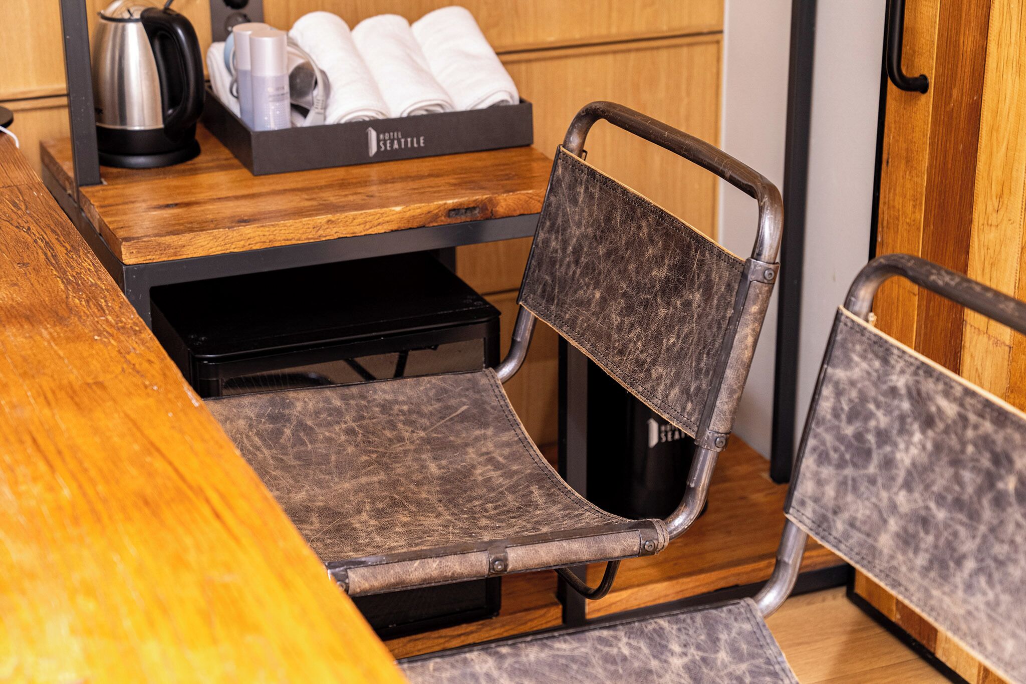 A wooden table with a black tray holding a kettle, cups, and neatly folded towels. A chair with a metal frame and leather seat is positioned at the table.