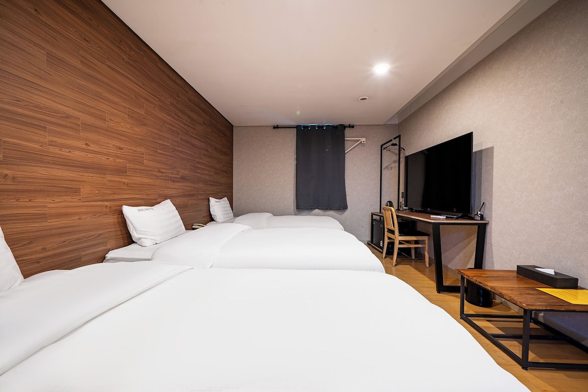 Ein modernes Hotelzimmer mit einer Wand in Holzoptik, einem Flachbildfernseher, einem Schreibtisch und einem Bett mit weißer Bettwäsche.