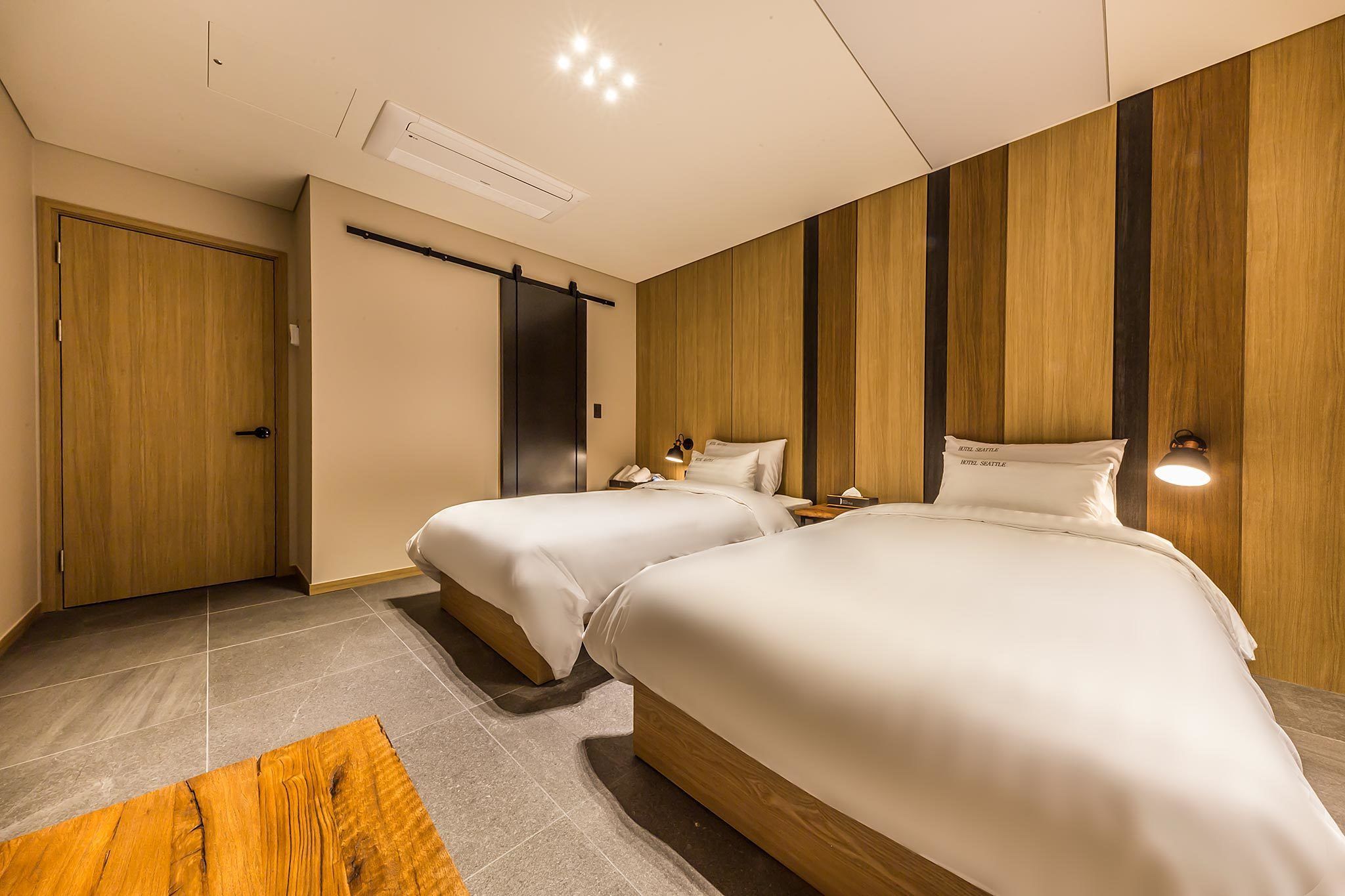 A hotel room with two beds, a wooden wardrobe, a desk, and a nightstand with a lamp.