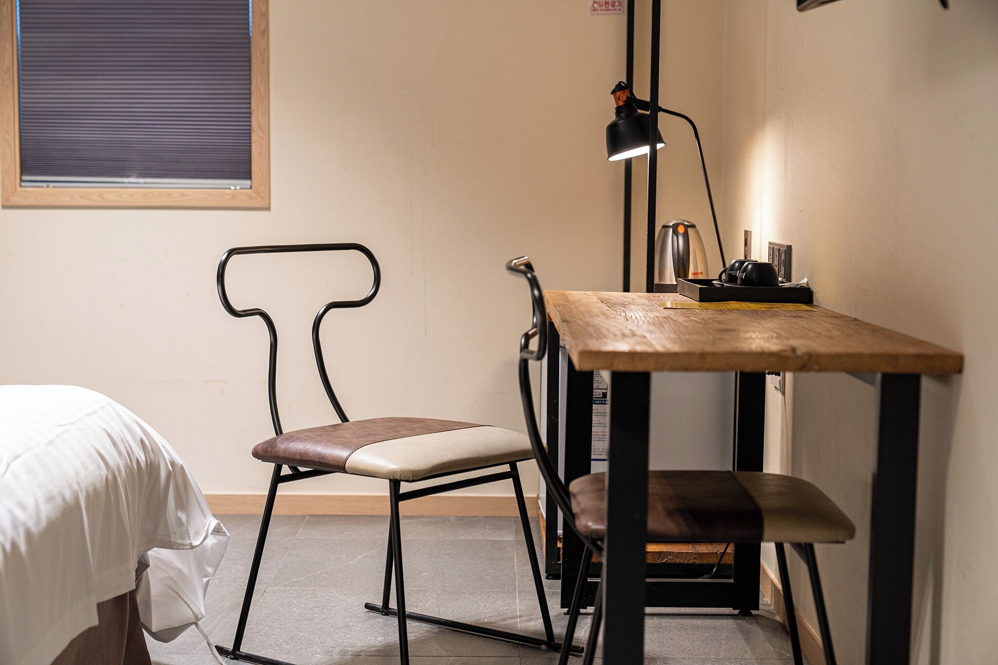 A small, cozy room with a bed, a wooden table, a chair, and a desk lamp.