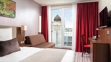 A hotel room with a large bed, a desk, and a chair. There is a window with red curtains and a view of a building with a dome.