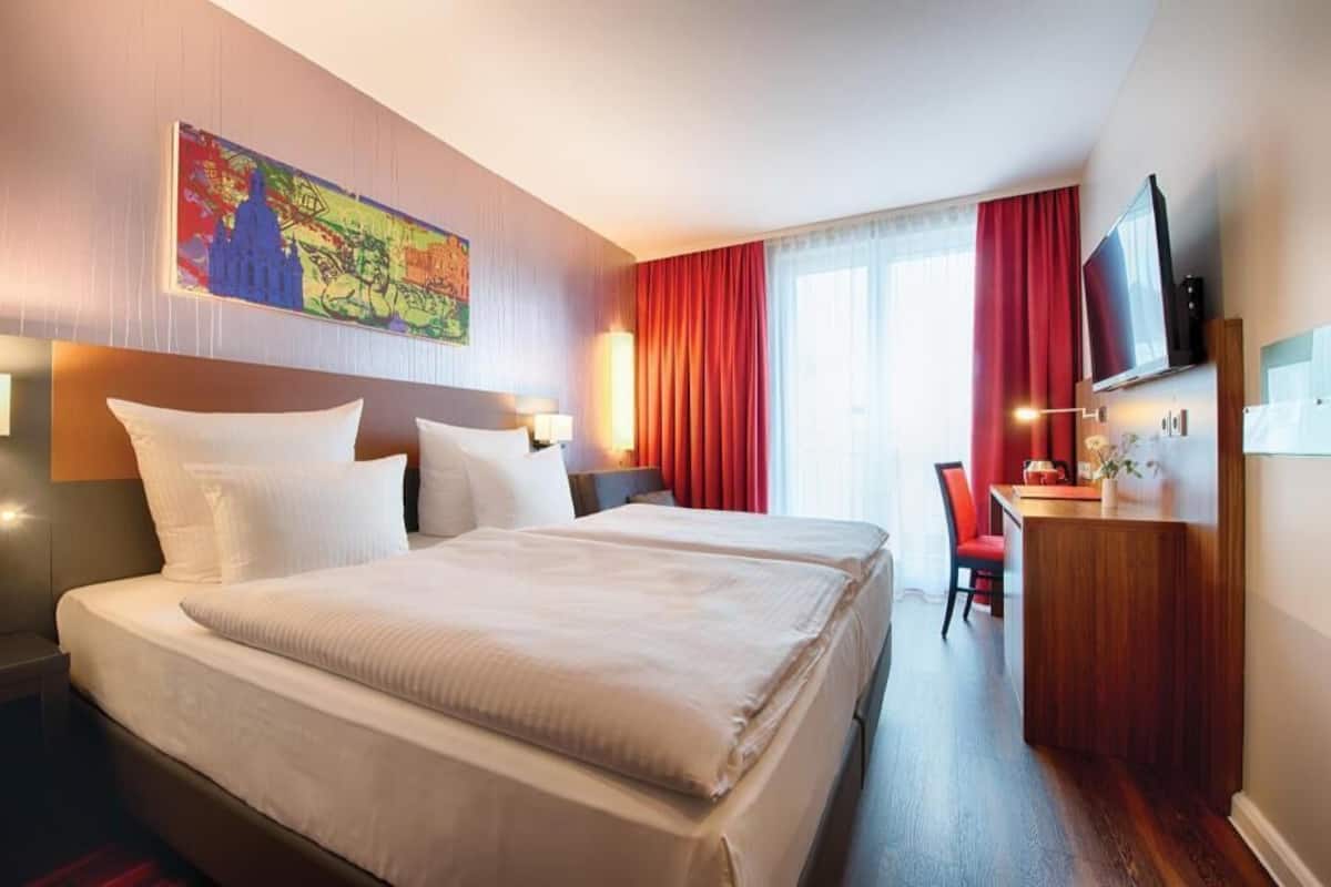 Quarto de hotel com uma cama grande, uma escrivaninha, televisão e um quadro na parede.