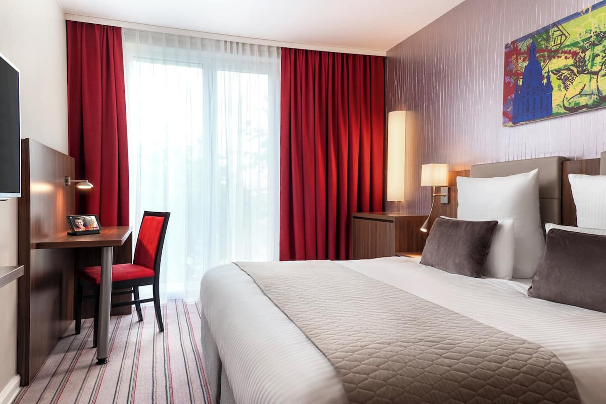 Quarto de hotel com uma cama grande, uma escrivaninha com cadeira, televisão e janela com cortinas vermelhas.