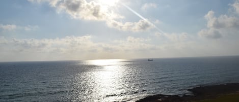 Uitzicht op de kust, met zonlicht dat op het water weerkaatst, een schip in de verte en een rotsachtige kustlijn.