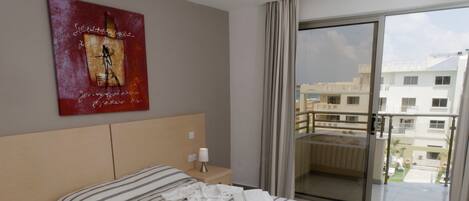 A hotel room with a bed, a nightstand, a painting on the wall, and a balcony with a view of buildings.