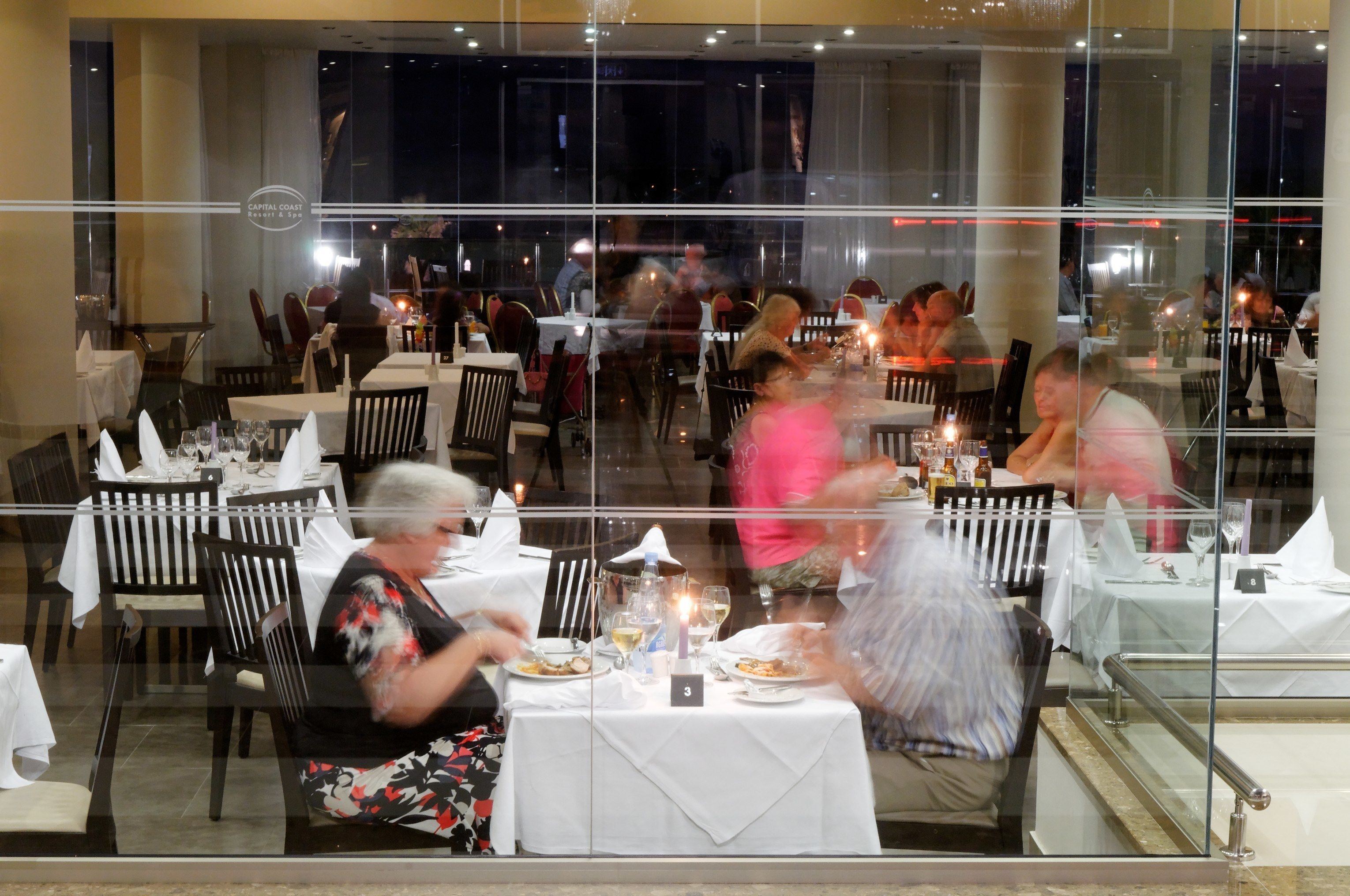 A restaurant with tables set for dining, a glass partition, and a view of the street outside.