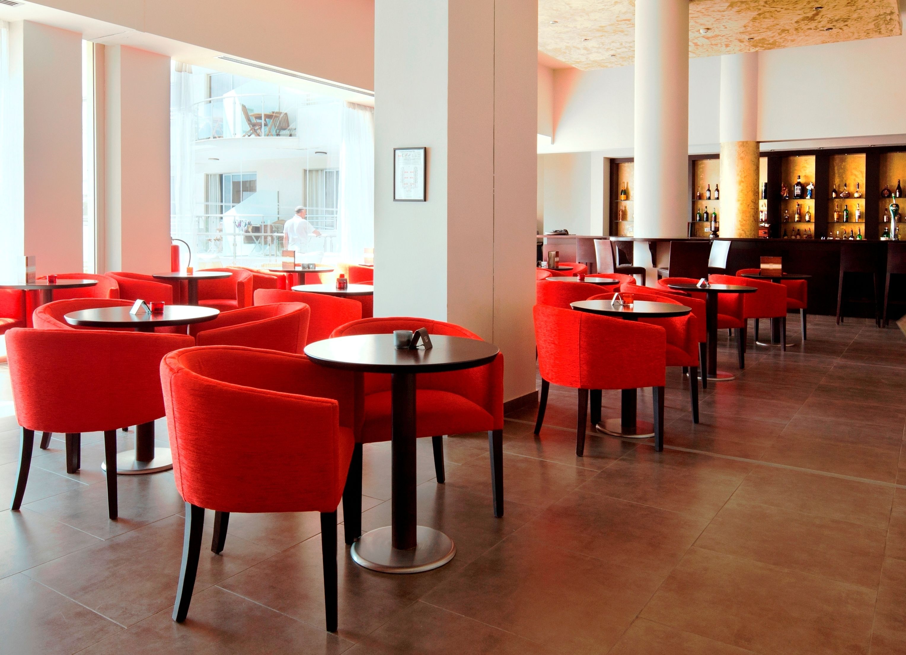 A modern restaurant with red chairs and round tables, a bar area with bottles, and a view of the outside.