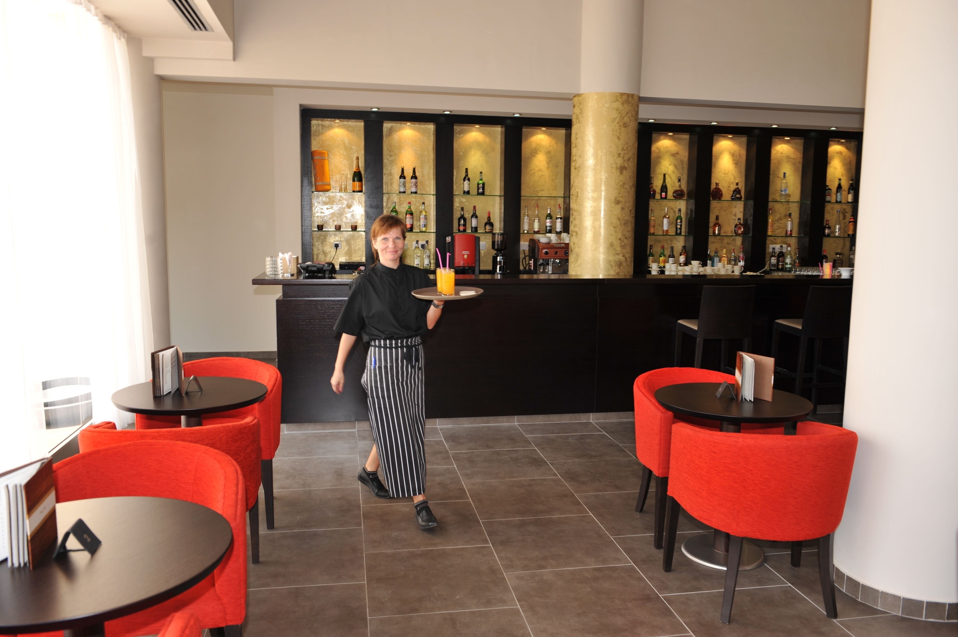 A bar area with a bartender carrying a tray, red chairs, and a bar counter with bottles.