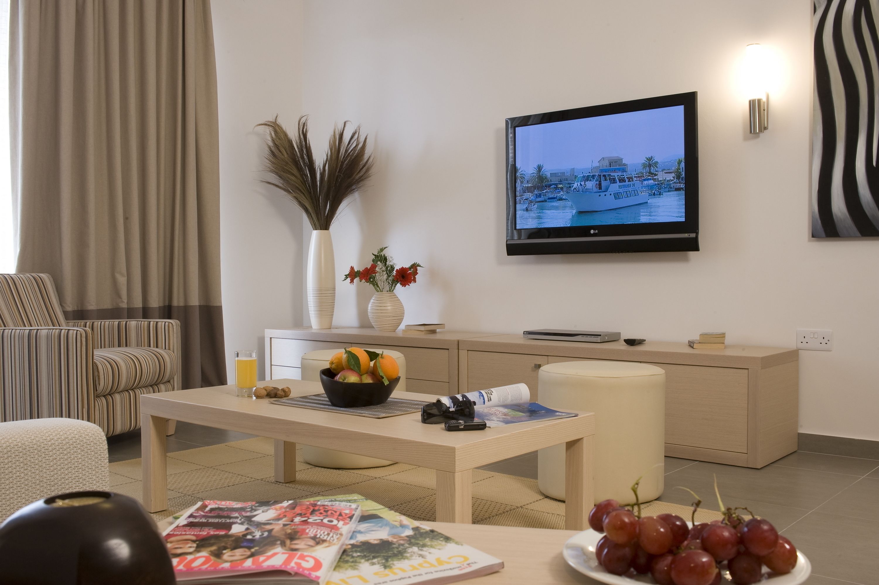 Uma sala de estar moderna com televisão de tela plana, uma mesa de jantar com frutas e um jornal, um sofá e um vaso com plantas secas.