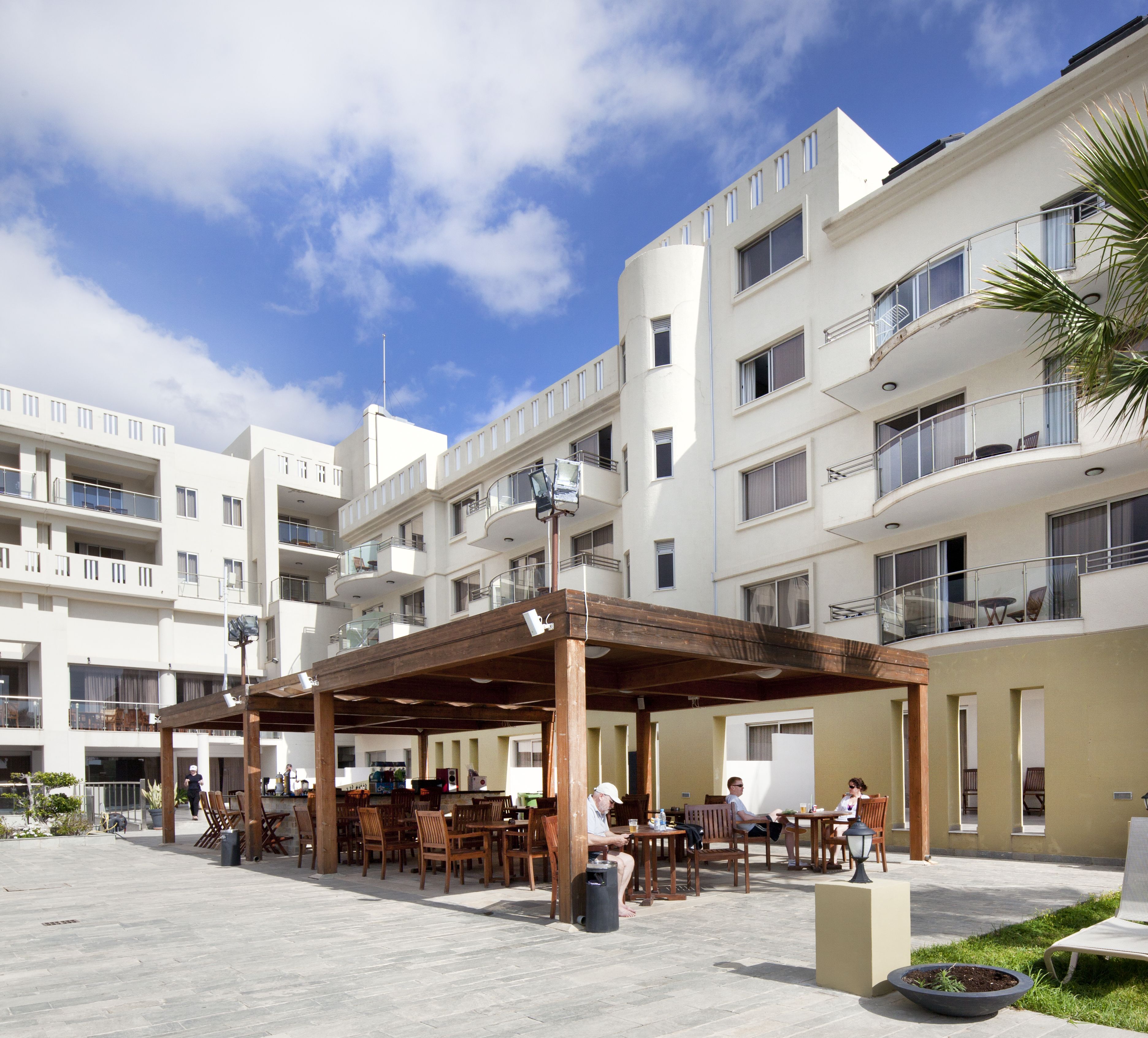 Un área de estar al aire libre cubierta, con mesas y sillas, rodeada por un edificio de varios pisos con balcones y ventanas.