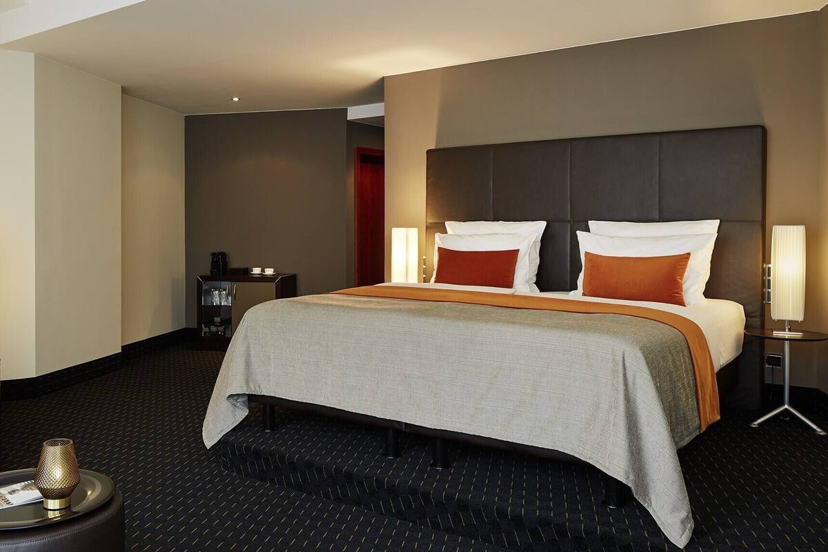 Habitación de hotel con una cama grande, una mesita de noche con lámpara, un armario pequeño y una mesa redonda con un objeto decorativo.