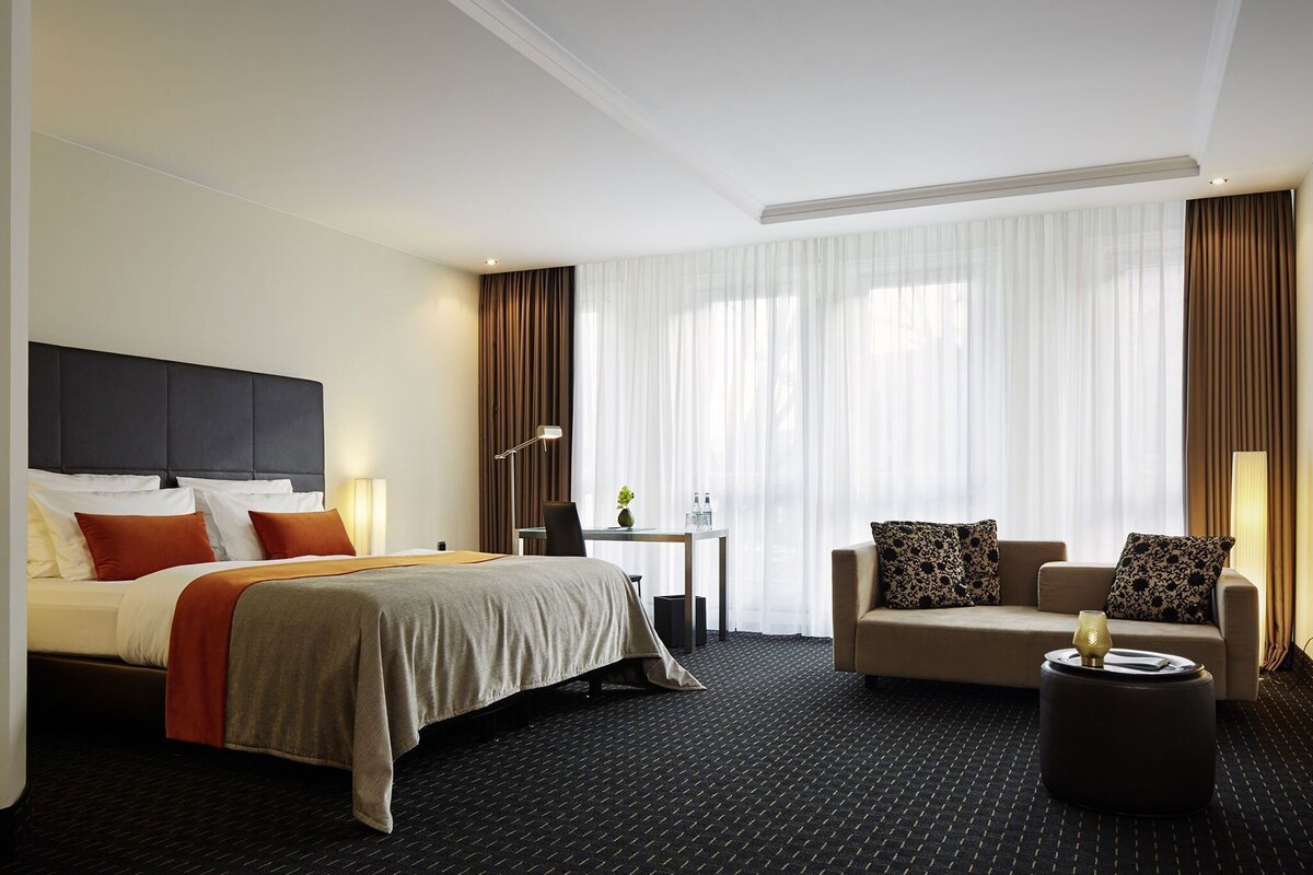 A hotel room with a bed, a sofa, a coffee table, a desk with a lamp, and a large window with curtains.