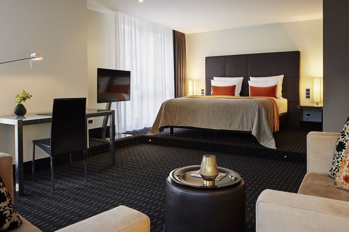 Habitación de hotel con cama, escritorio con silla, televisor, lámpara y una pequeña mesa con un objeto decorativo.