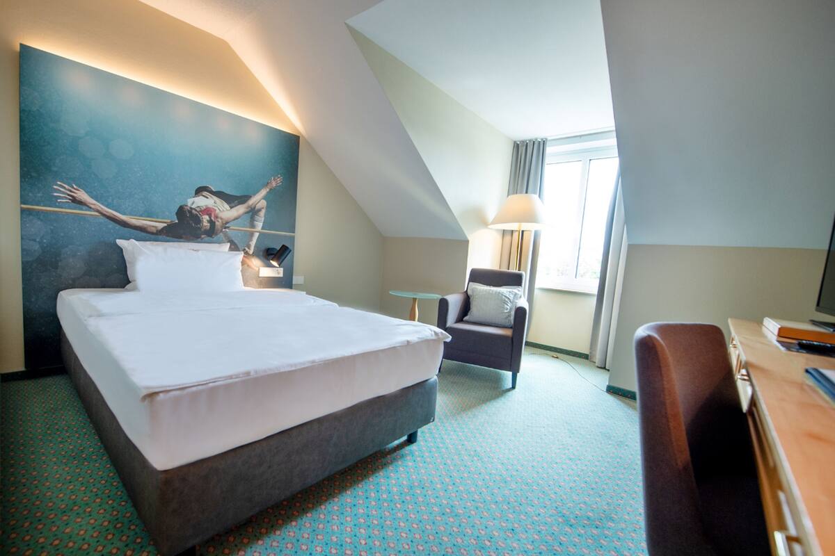 A hotel room with a large bed, a desk, a chair, a lamp, and a painting on the wall.