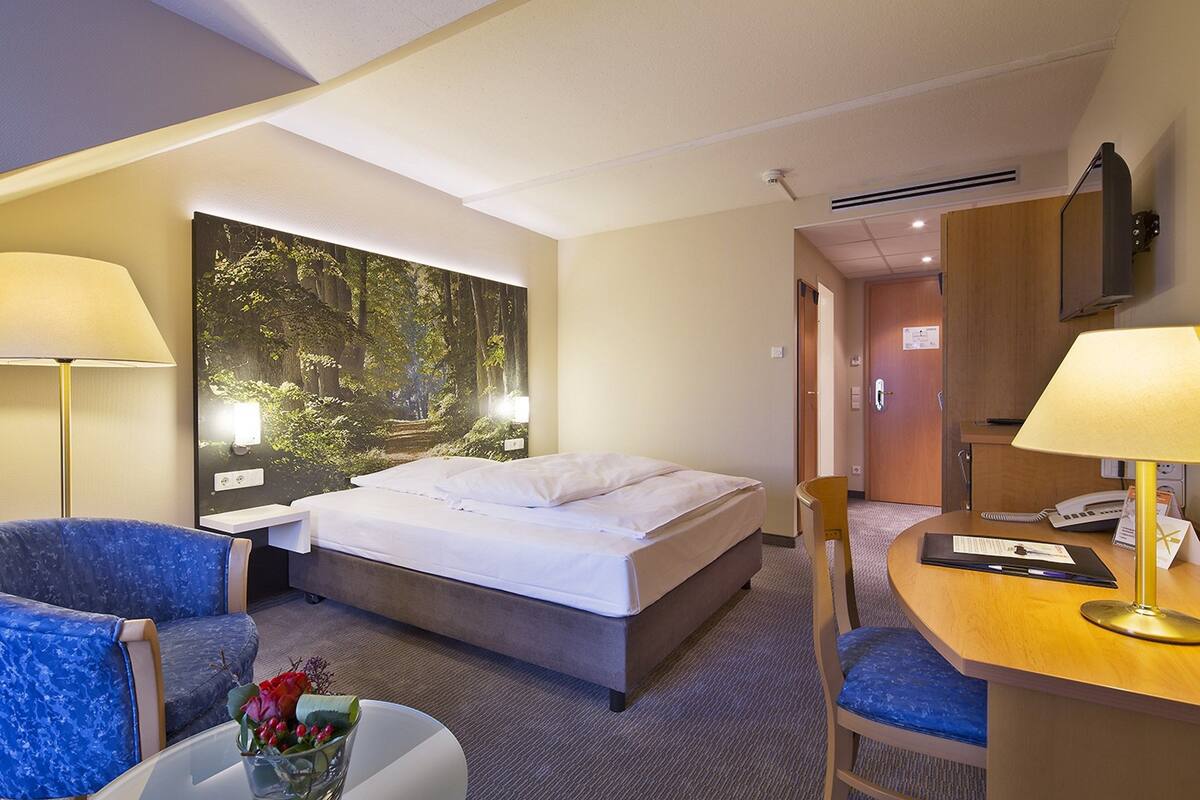 A hotel room with a bed, two chairs, a desk with a lamp, a wall-mounted TV, and a large forest-themed painting.
