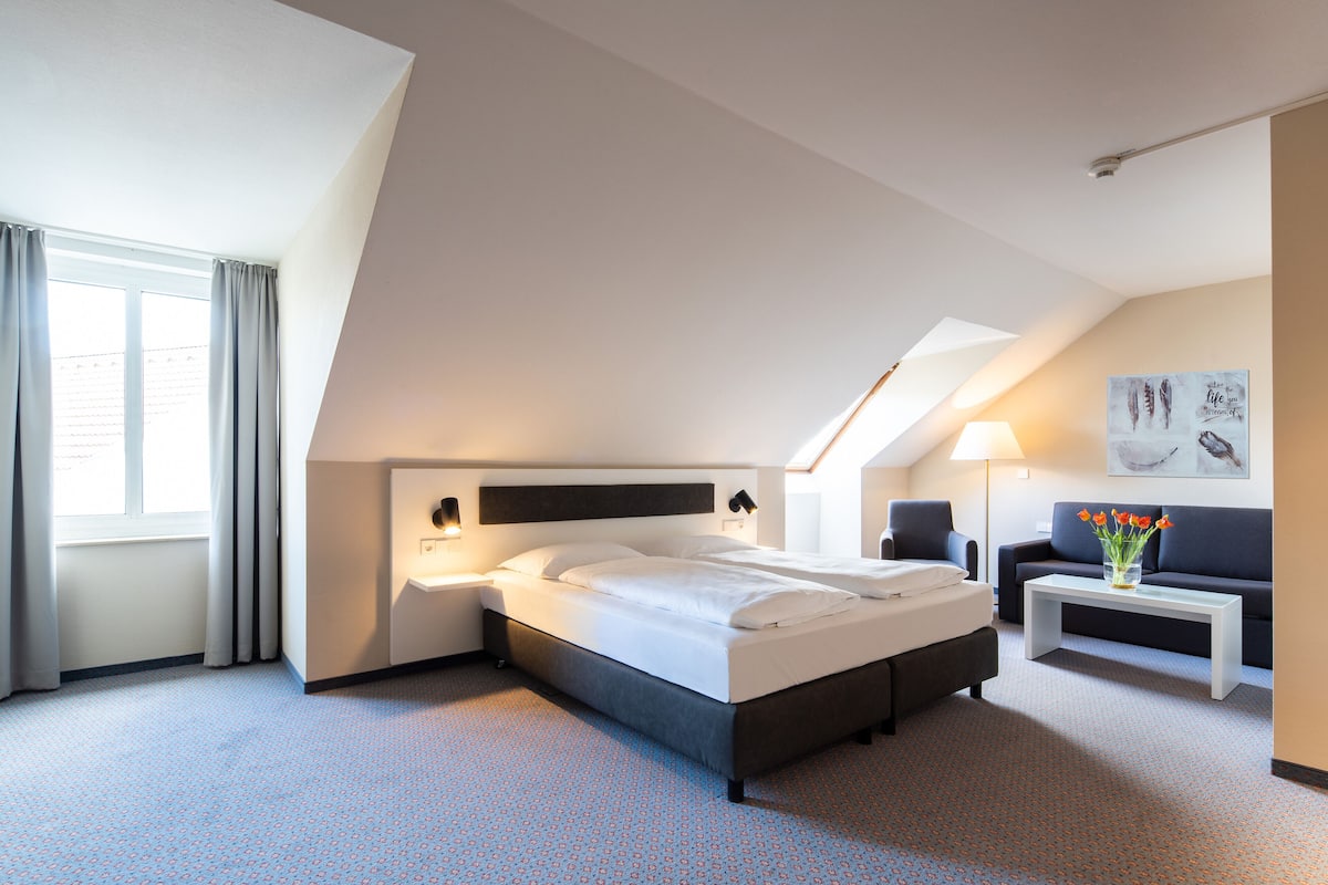 A modern hotel room with a large bed, a small sofa, a nightstand, and a lamp.