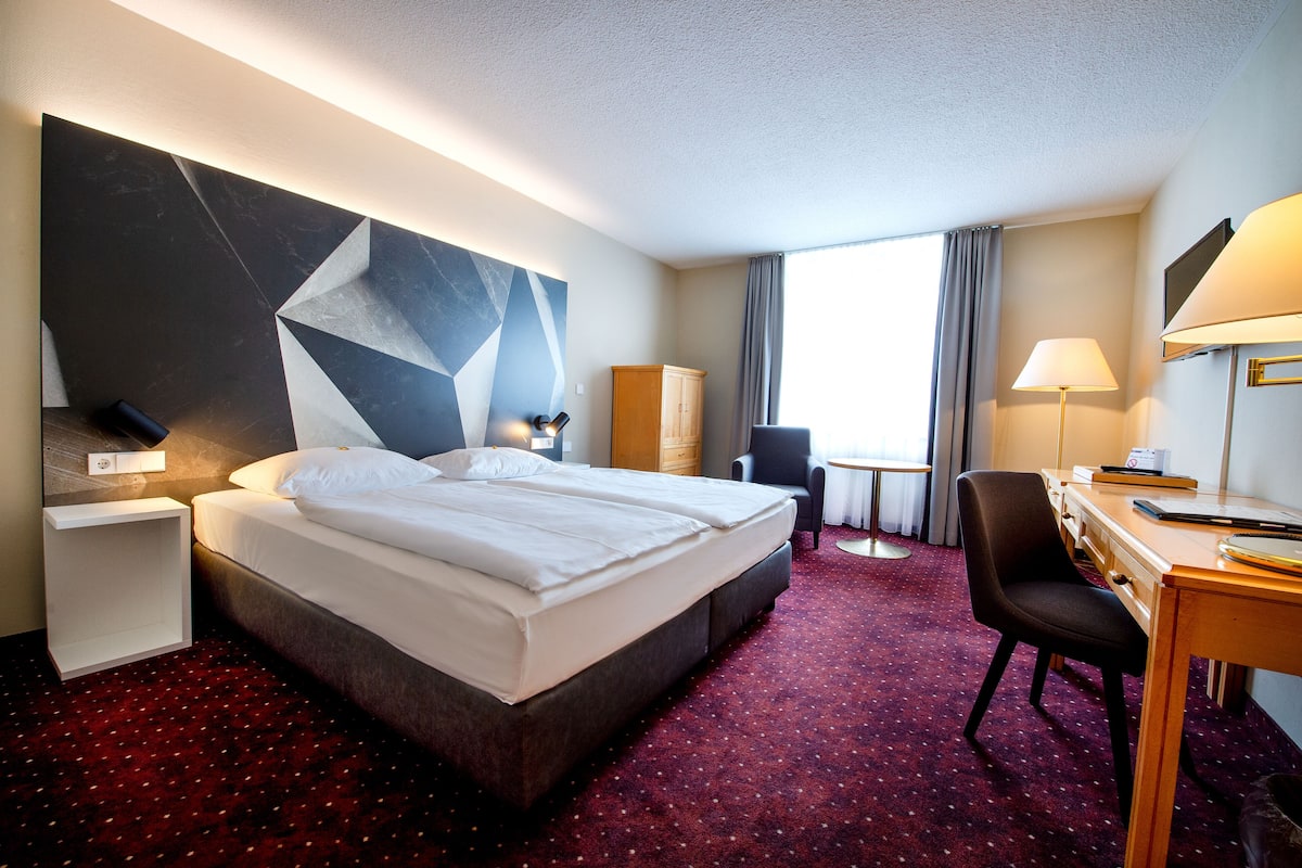 A hotel room with a large bed, a desk, a chair, a small table, a wardrobe, and a TV.