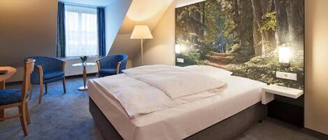 A hotel room with a bed, a desk, a chair, a lamp, and a large forest-themed wall mural.