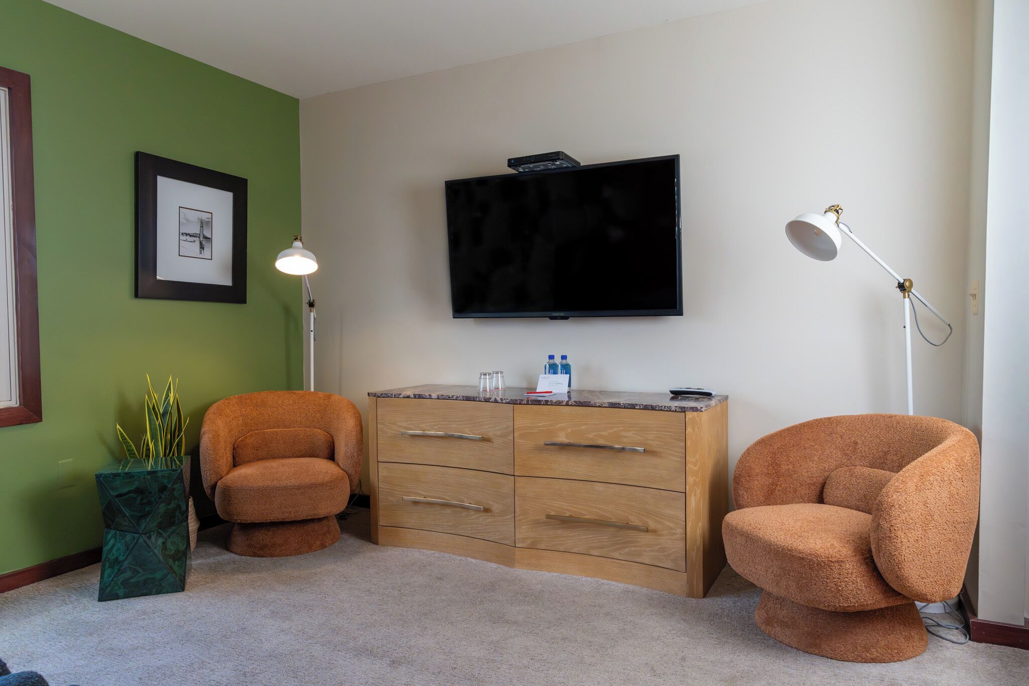 A room with a flat-screen TV mounted on the wall, a wooden cabinet with drawers, two orange armchairs, a floor lamp, and a framed picture on the wall.