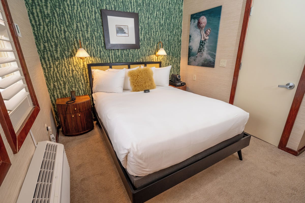 A neatly made bed with white linens, a headboard with a light fixture, a bedside table with a lamp, and a wall with a framed picture.