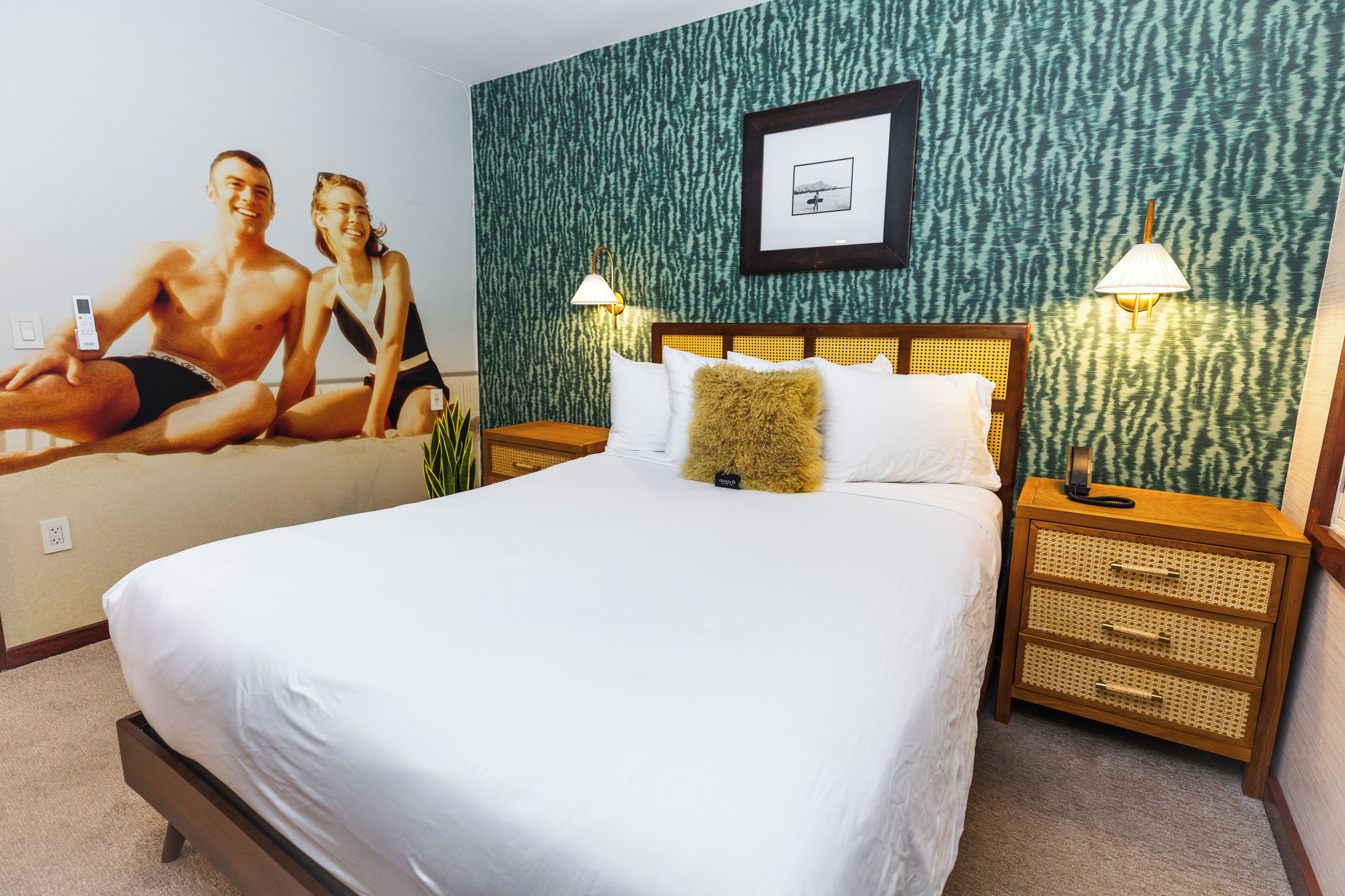 Habitación de hotel con una cama grande, dos mesitas de noche con lámparas, un cuadro enmarcado en la pared y una pared verde con textura.