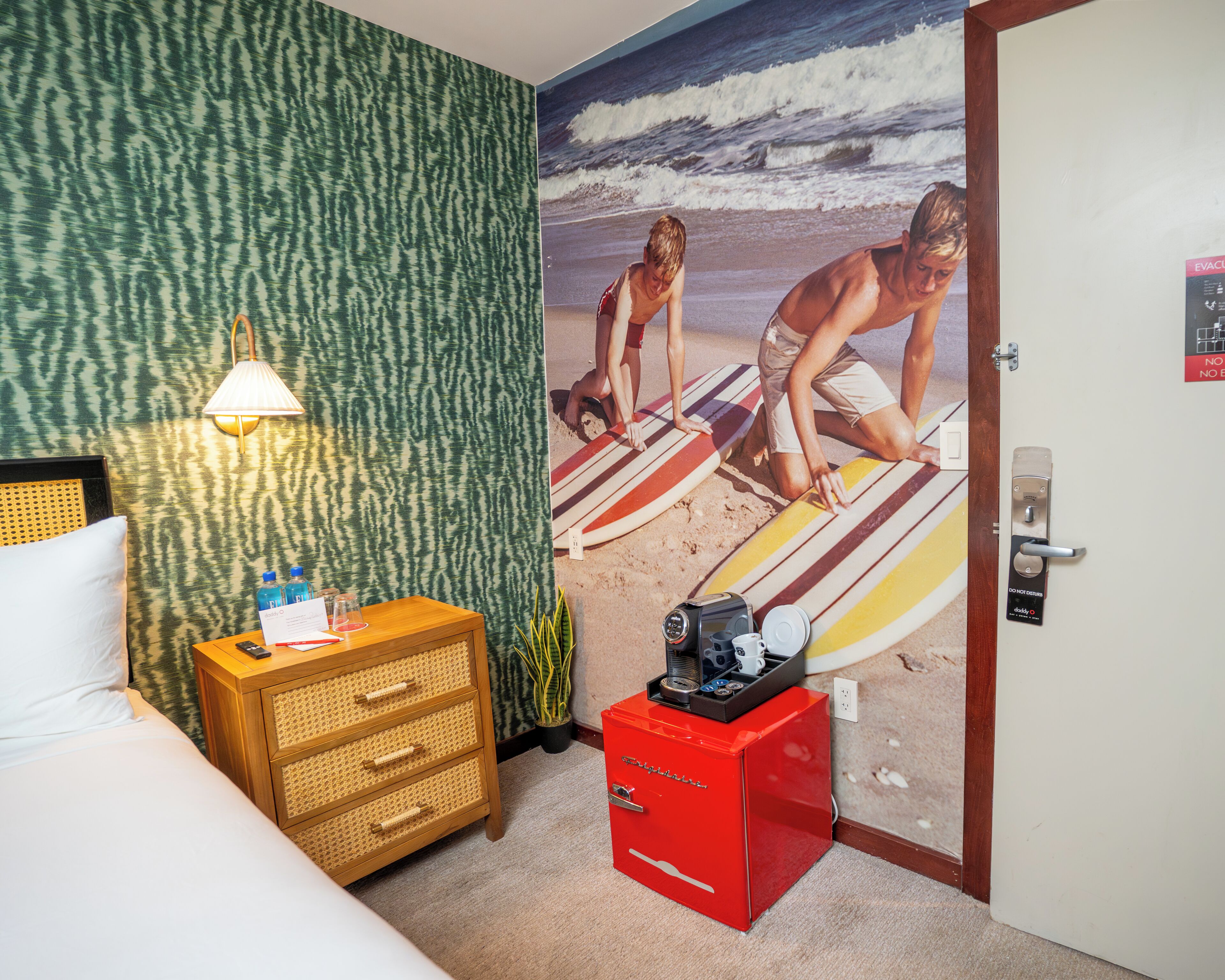 Habitación de hotel con cama, mesita de noche con lámpara, minibar rojo, pared con un mural de playa y puerta con letrero de salida de emergencia.