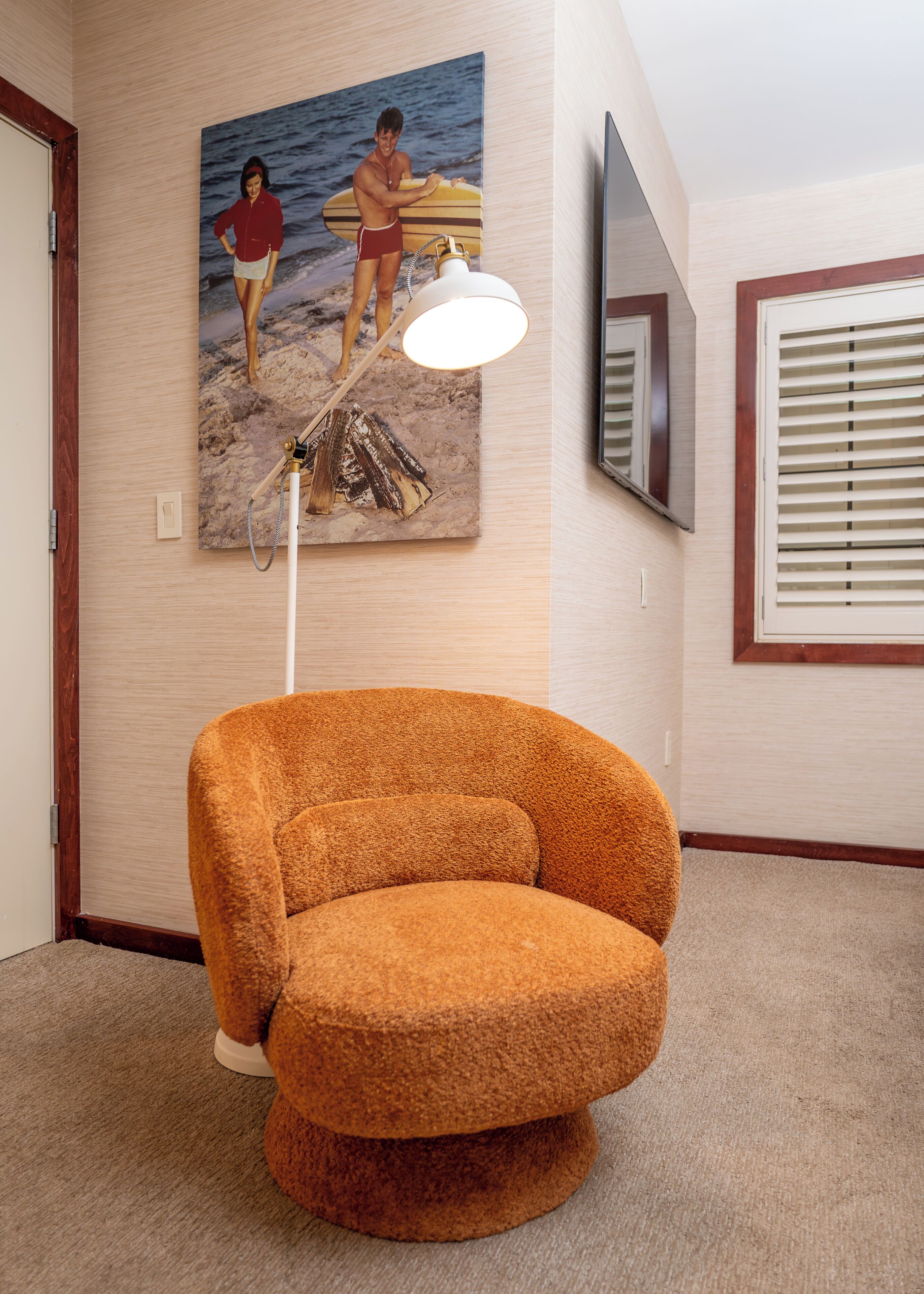 A room with a round orange chair, a painting of a beach scene, a ceiling-mounted lamp, and a TV on the wall.