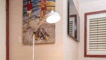 A room with a round orange chair, a painting of a beach scene, a ceiling-mounted lamp, and a TV on the wall.