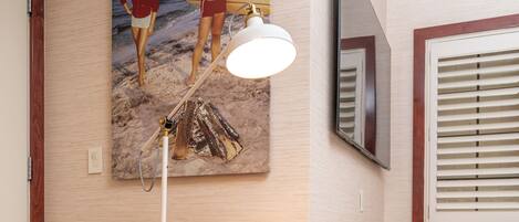 A room with a round orange chair, a painting of a beach scene, a ceiling-mounted lamp, and a TV on the wall.