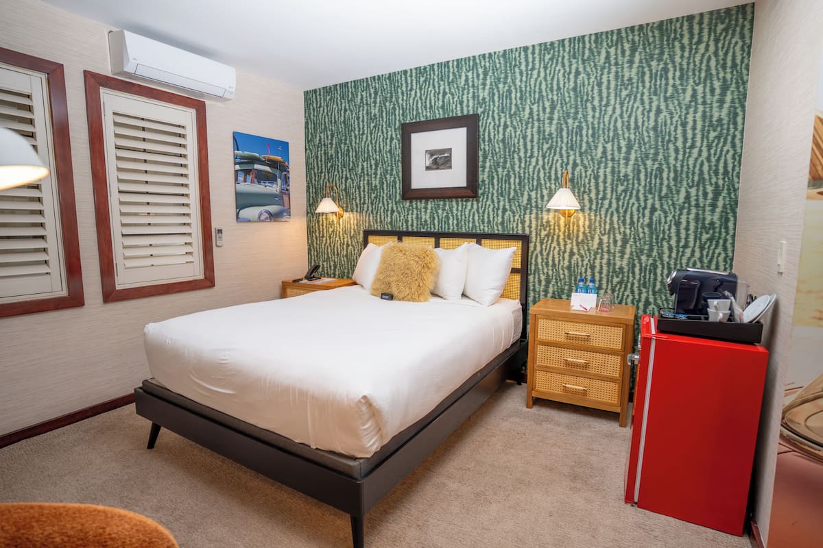 A hotel room with a bed, bedside lamps, a small wooden nightstand, a red refrigerator, and a wall-mounted air conditioner.