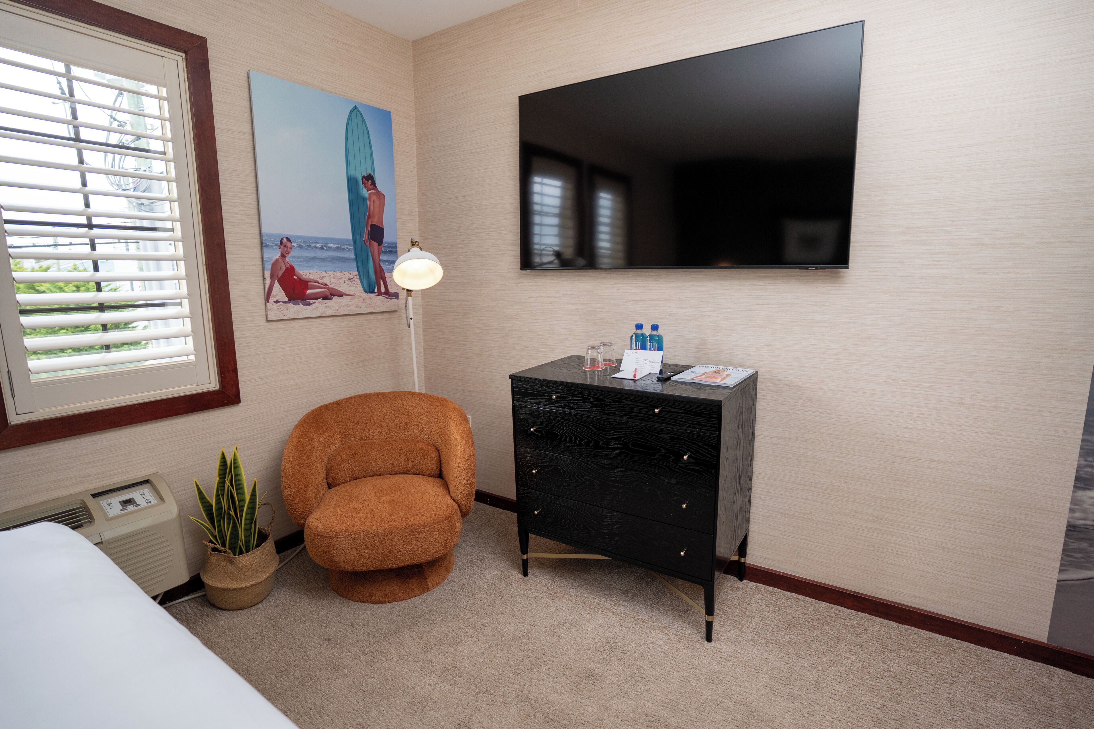 Une chambre d’hôtel avec un lit, un fauteuil marron, une commode noire, un téléviseur à écran plat, un tableau encadré, une lampe et une fenêtre avec des stores.